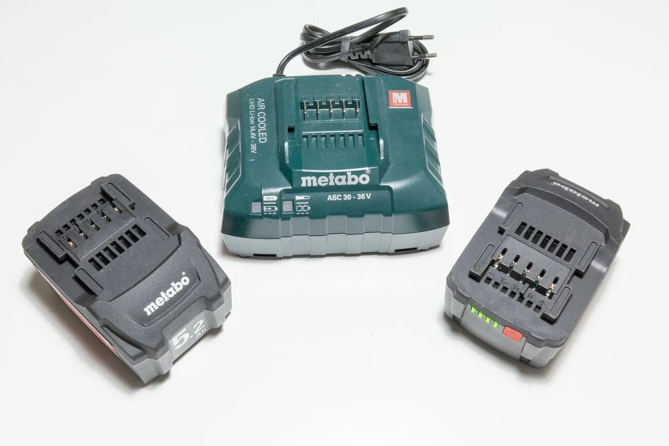 Zwei schwarze Akkus für den Metabo STA 18 LTX 140 liegen auf einer weißen Oberfläche. Dazwischen liegt das grüne Metabo Ladegerät für die Akkus.